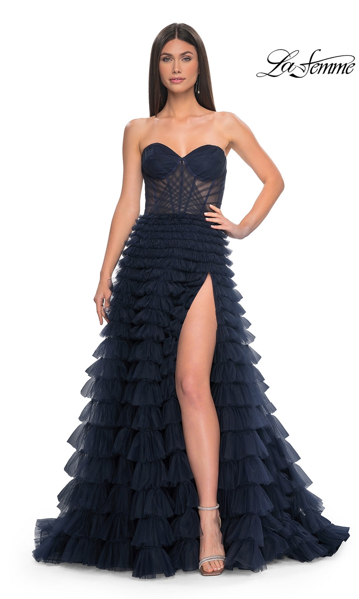 A woman poses confidently with one hand on her hip in the La Femme Strapless Long Ruffled Prom Dress 32283, a navy blue A-line gown featuring a sheer corset bodice and high leg slit, set against a white background.